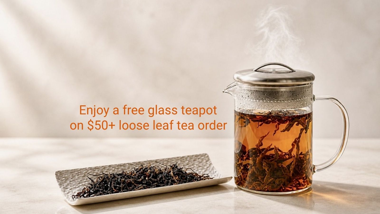 A clear glass teapot filled with amber tea sits on a light surface beside a tray of loose leaf tea, with orange text reading 'Enjoy a free glass teapot on $50+ loose leaf tea order' against a soft beige background.
