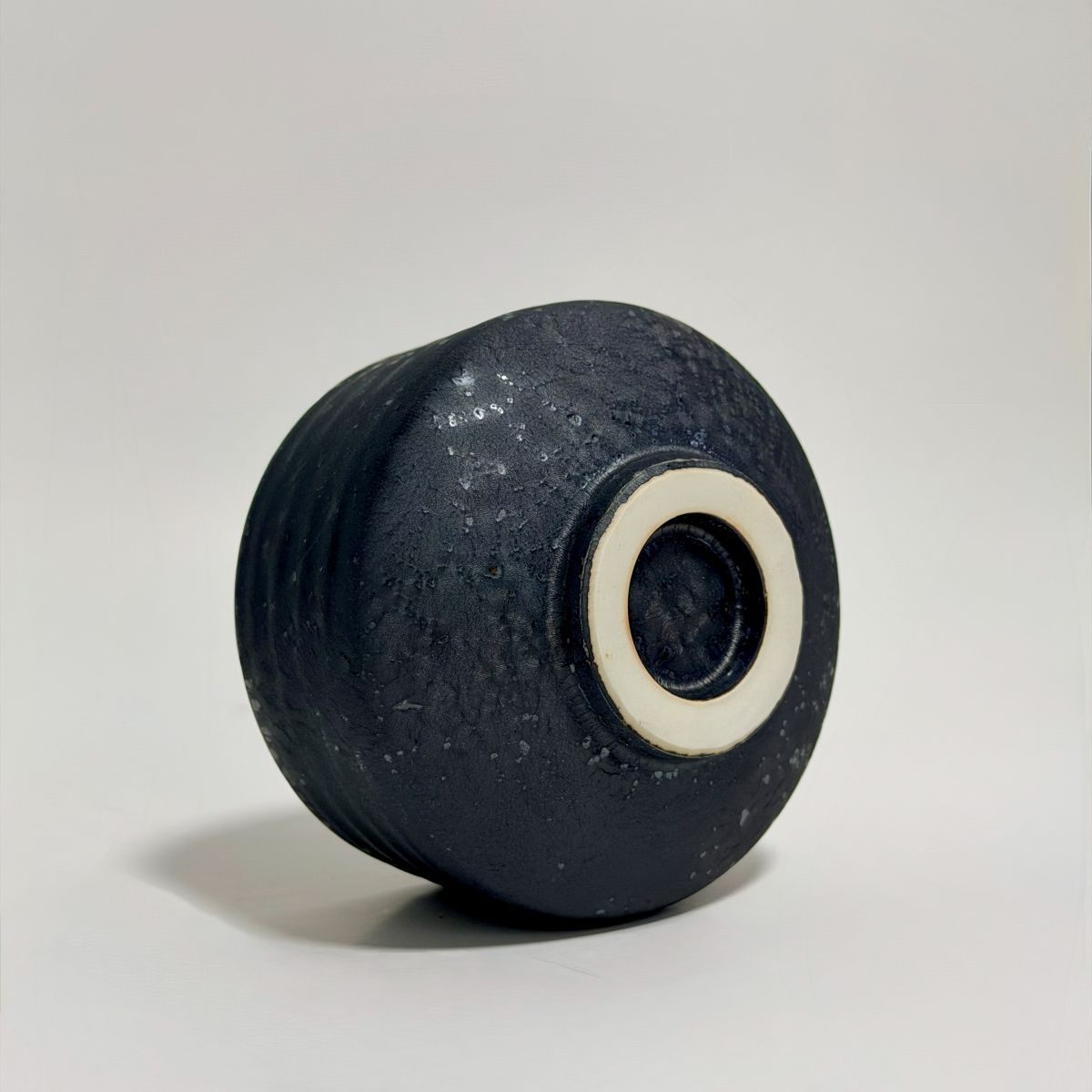 A dark textured ceramic vase with a ribbed body and a contrasting off-white ring around the base, photographed on a neutral light gray background.