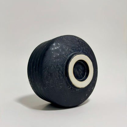A dark textured ceramic vase with a ribbed body and a contrasting off-white ring around the base, photographed on a neutral light gray background.