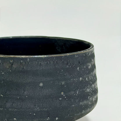 Close-up of a dark charcoal gray ceramic bowl with a textured, speckled glaze and a smooth rim, set against a plain light gray background.