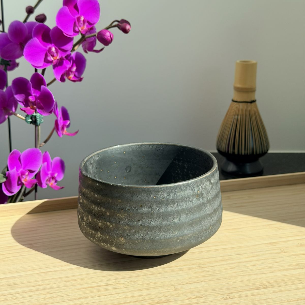 A dark gray textured ceramic matcha bowl with horizontal ridges sits on a light wood table, with purple orchids on the left and a bamboo whisk in the background.