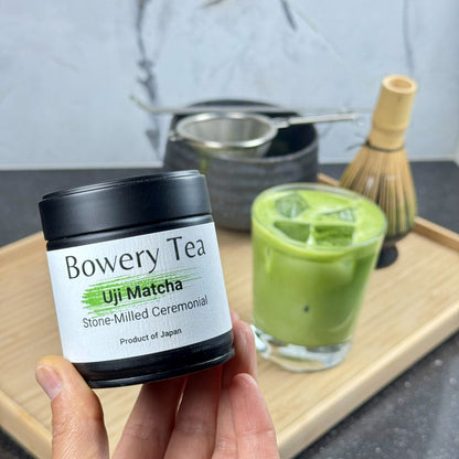Bowery Tea Uji Matcha container held by a hand with a glass of matcha in the background.