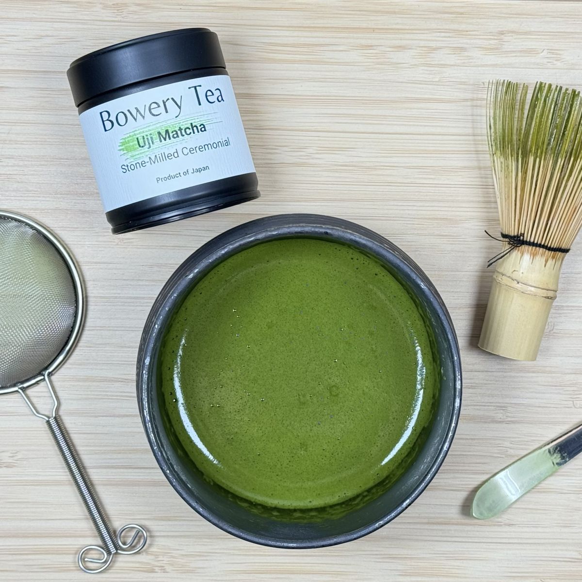 Bowery Tea Uji Matcha with a bowl of green tea powder, whisk, and other tools on a wooden surface.