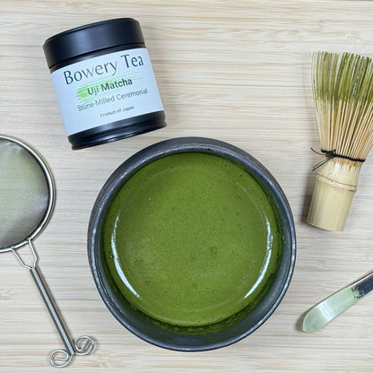 Bowery Tea Uji Matcha with a bowl of green tea powder, whisk, and other tools on a wooden surface.