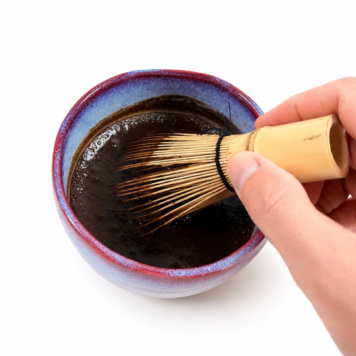 Hand using a bamboo whisk to stir dark liquid in a ceramic bowl on a white background