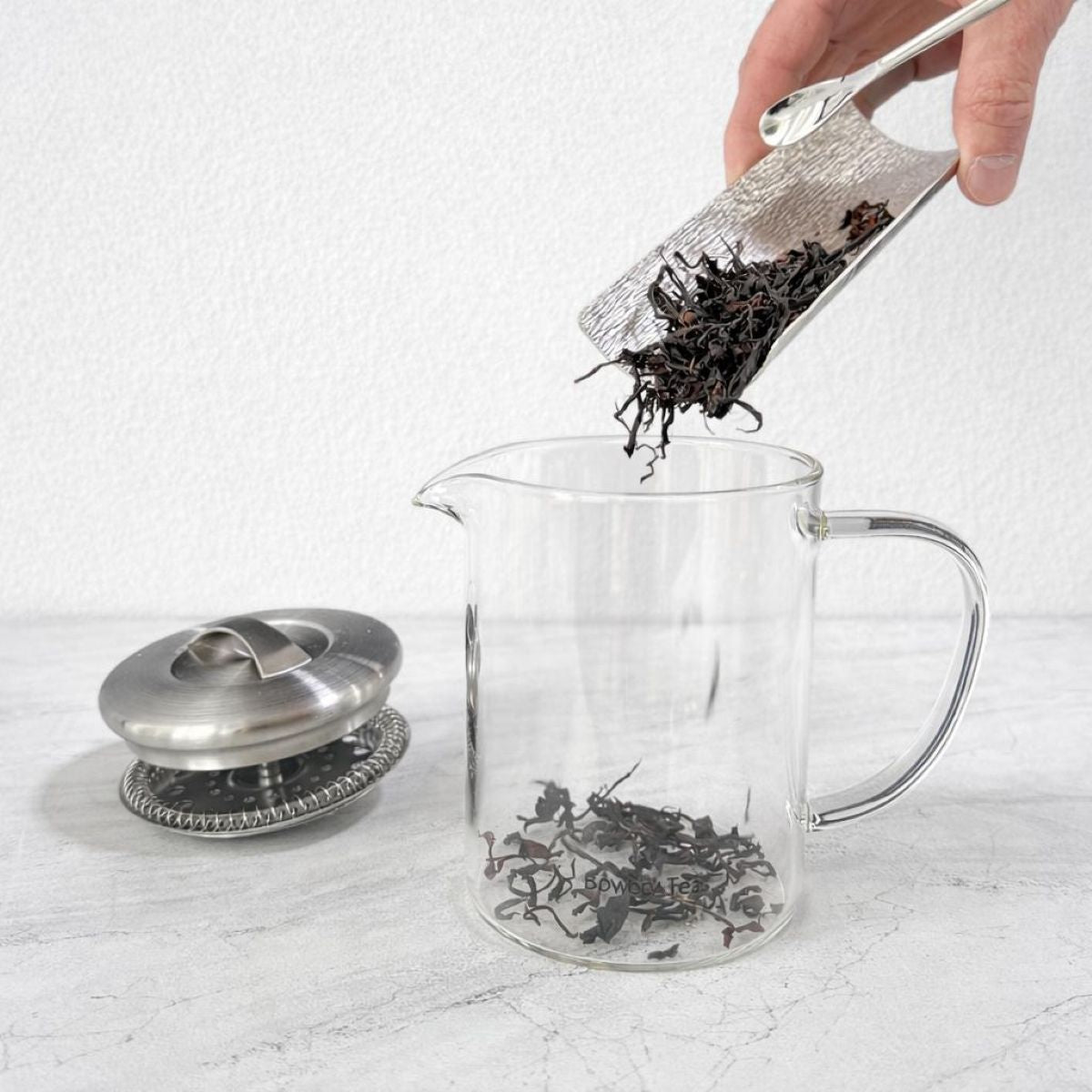 Loose leaf tea being added to a glass teapot with an infuser for brewing