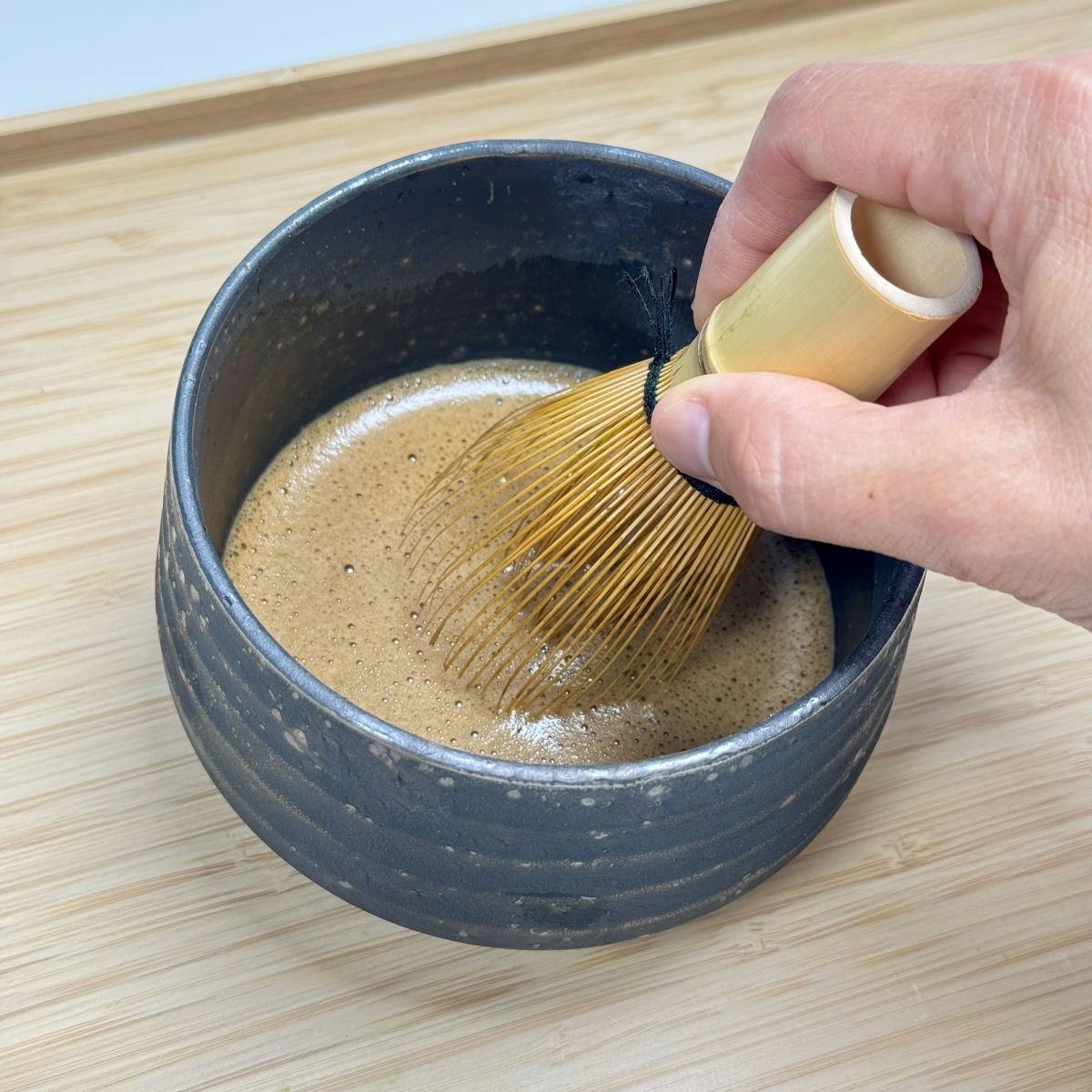 Person using a bamboo whisk to stir matcha in a black ceramic bowl on a wooden surface.